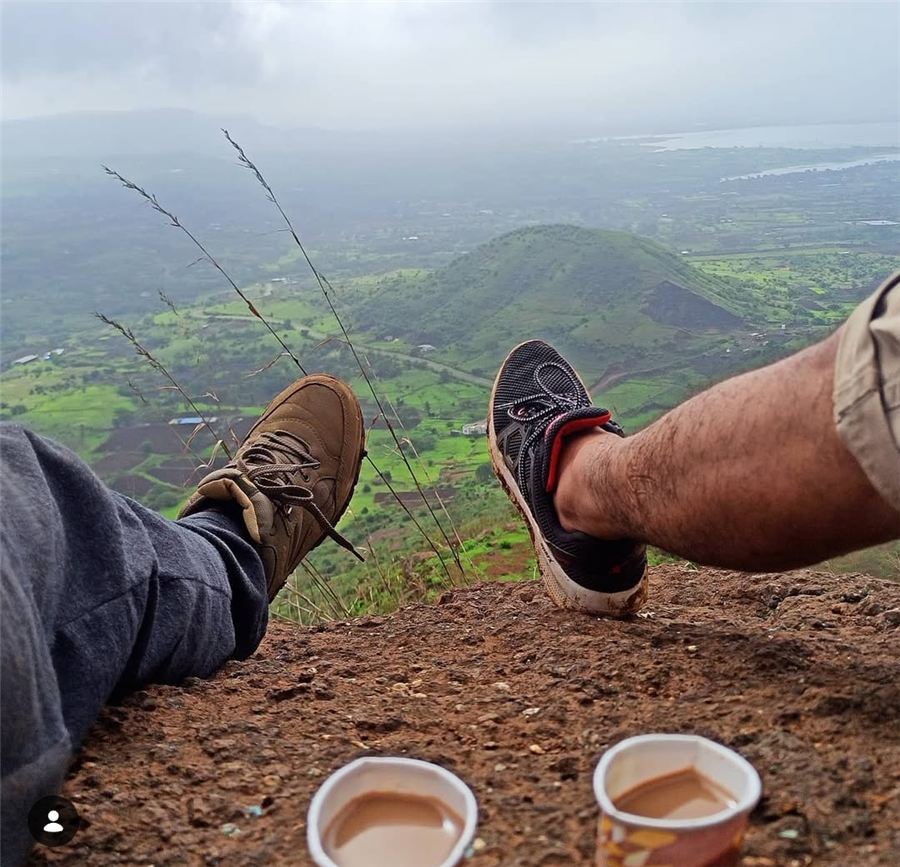 Two pairs of trekking shoes and cups of chai on a cliff above a green valley