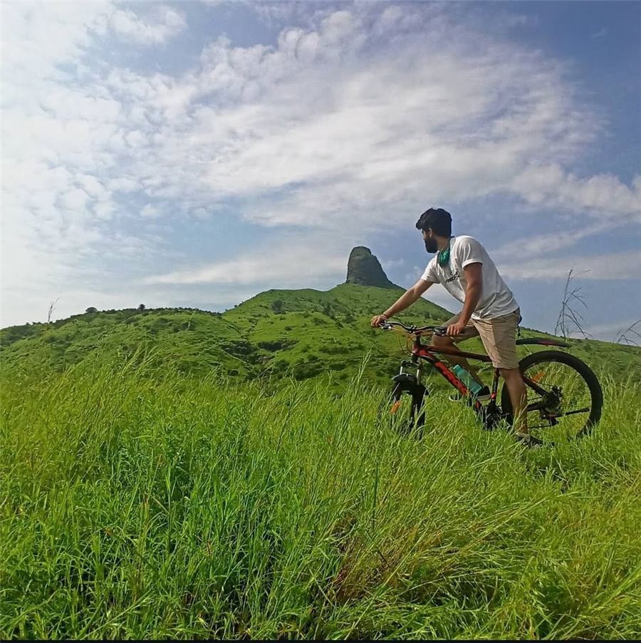 Pratham on a bicycle in tall green grass, looking toward a hill