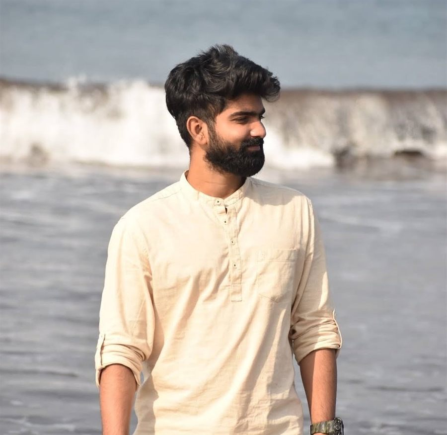 Pratham standing on a beach in a cream kurta, waves breaking behind him
