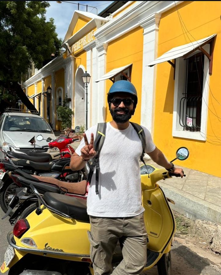 Pratham giving a peace sign beside a yellow moped parked on a street of yellow colonial buildings